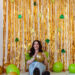 A model in front of a gold fringe background that has clovers attached to it that are green. The model has on a lime green top, light wash jeans, and white dr marten boots. She has brown hair and is holding a gold balloon. There are gold coins, lime green balloons, and gold balloons strewn around her.